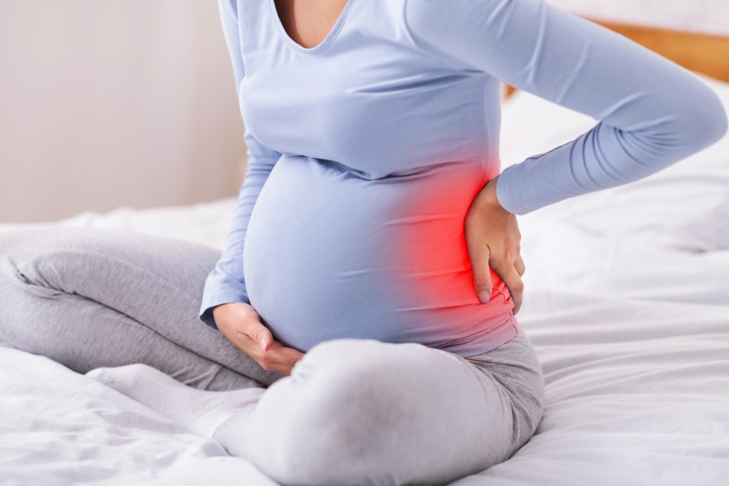 Pregnant woman sitting on bed with back pain, our St. Petersburg Chiropractor can administer prenatal adjustments.